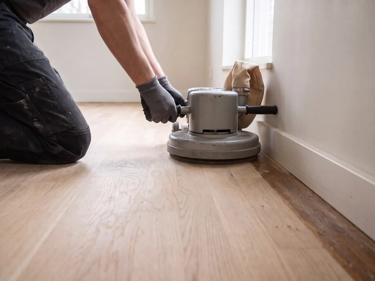 Hände in Arbeitshandschuhen führen eine Randschleifmaschine an der Sockelleiste entlang; an der Wand ist noch ein schmaler alter, dunkler Streifen