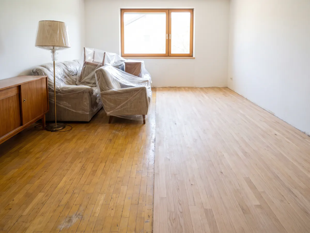 Wohnzimmer mit zusammengerückten, in Folie gehüllten Möbeln auf dem alten Parkett; die andere Raumhälfte ist schon frisch geschliffen